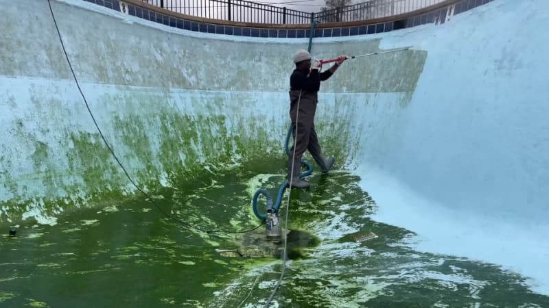 Technician washing algae from the sloping wall of a drained swimming pool