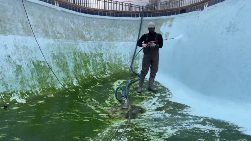 Technician working a pressure washer across the algae-streaked wall of a drained pool