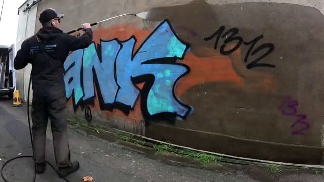 Technician using a pressure washer to strip spray-painted graffiti from a concrete wall