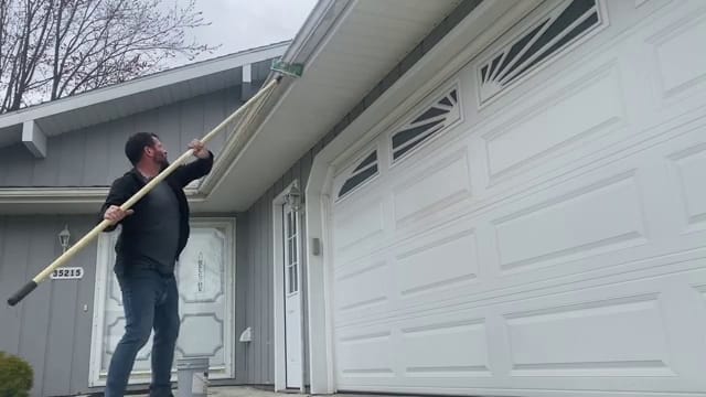 Technician scrubbing the face of a gutter above a garage door with a long extension pole