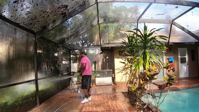 Technician washing a pool cage screen from inside the enclosure, beside a brick paver deck