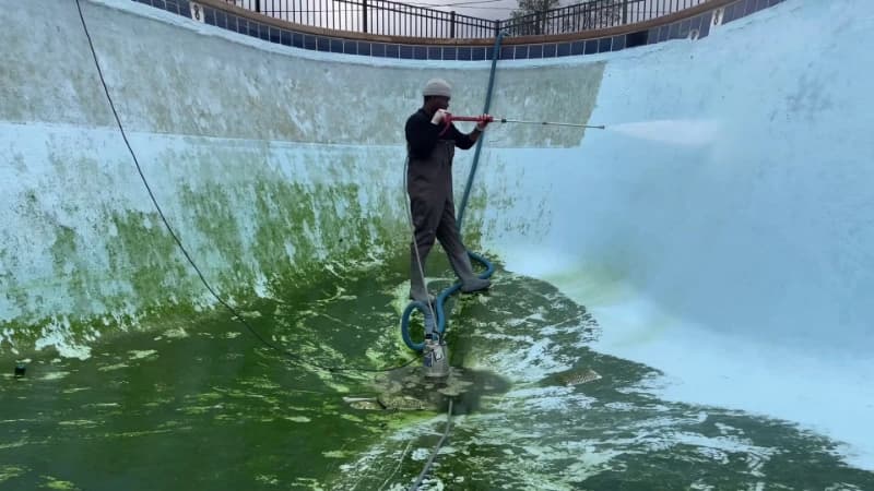 Technician washing green algae from the curved wall of a drained swimming pool