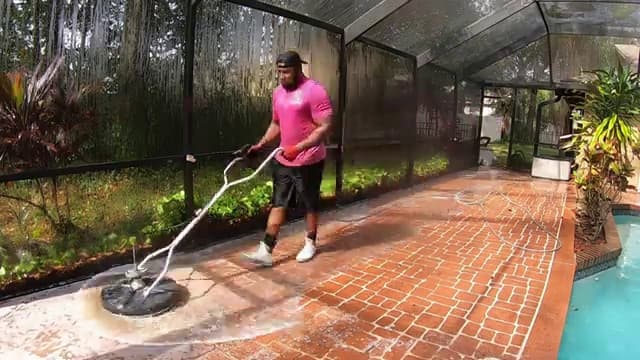 Technician running a surface cleaner across a brick paver pool deck inside a screen enclosure
