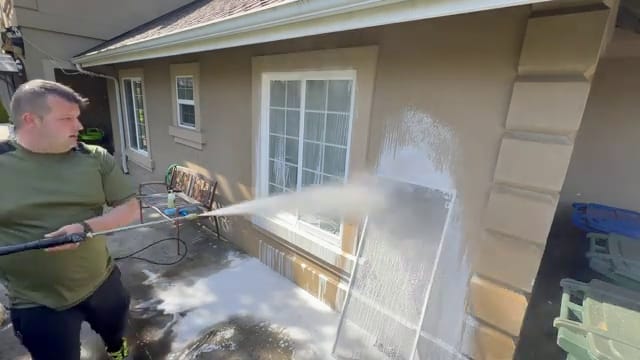 Technician rinsing a removed window screen propped against the stucco wall of a home