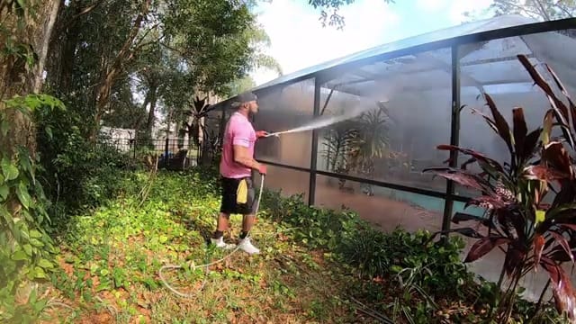 Technician rinsing the outside of a screened pool enclosure, with the pool visible through the mesh